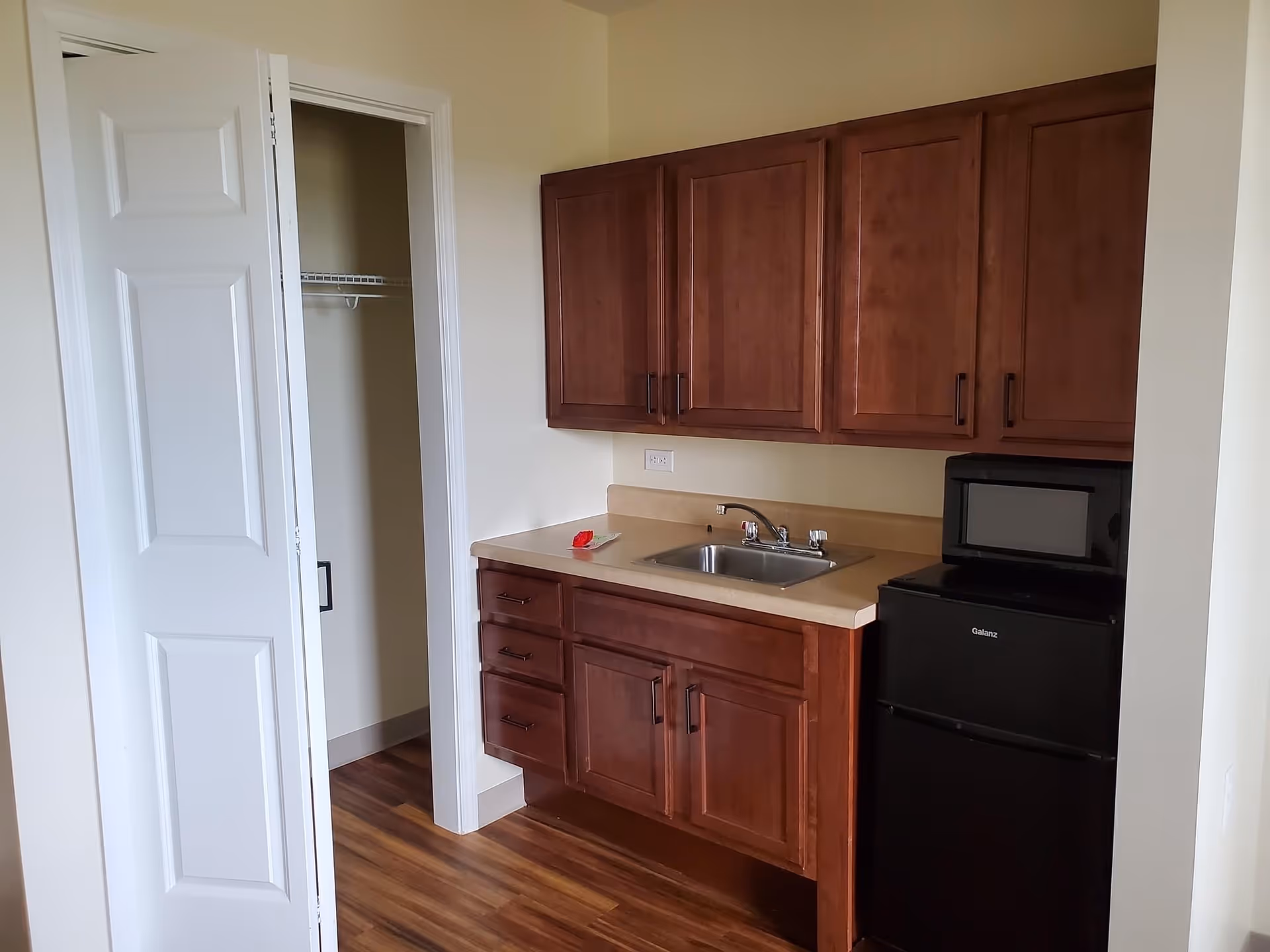 Small kitchenette area with wooden cabinets above and below a countertop with a sink. To the right, there is a black microwave on top of a black mini refrigerator. To the left, there is an open door leading to a closet with a wire shelf. The floor is wood and the walls are light-colored.