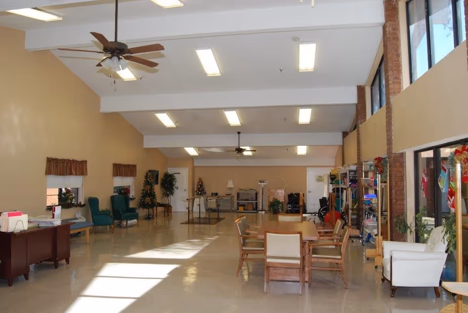 A spacious room with beige walls and a high ceiling featuring ceiling fans and fluorescent lights. The room has large windows on one side letting in natural light. There are several chairs and tables arranged in the room, including a wooden table with chairs in the center and green armchairs along the wall. The room also contains some plants, shelves with various items, and a desk with papers and folders. The overall setting appears to be a common area or lounge in a senior living facility.