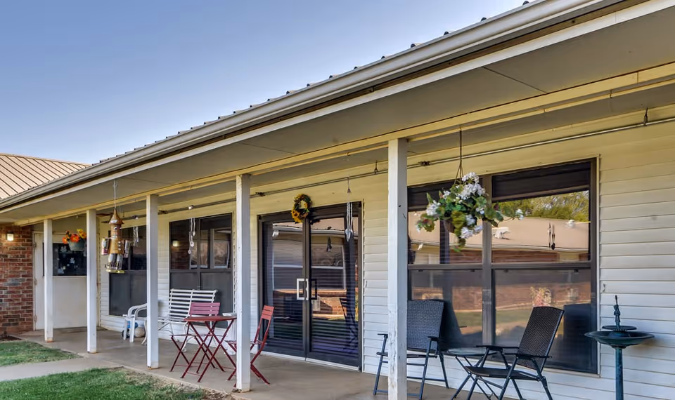 Covered outdoor patio area at Pleasant Valley facility with white pillars, hanging flower baskets, wind chimes, and outdoor seating including a small red table with two chairs and two black chairs with a small round table. The building exterior is light-colored siding with large windows and a glass door.