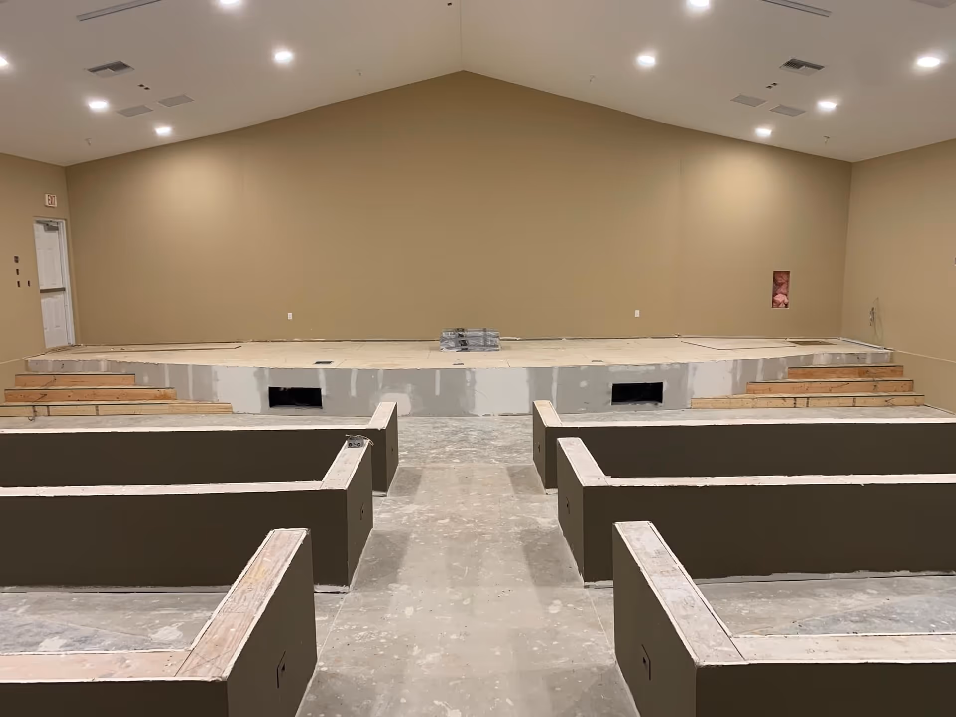 An unfinished interior room with a raised stage at the far end and several rectangular wooden frame structures on the floor, possibly for seating or partitions. The walls are painted beige, and the ceiling has multiple recessed lights. The floor is bare concrete, and there are stairs on either side of the stage.