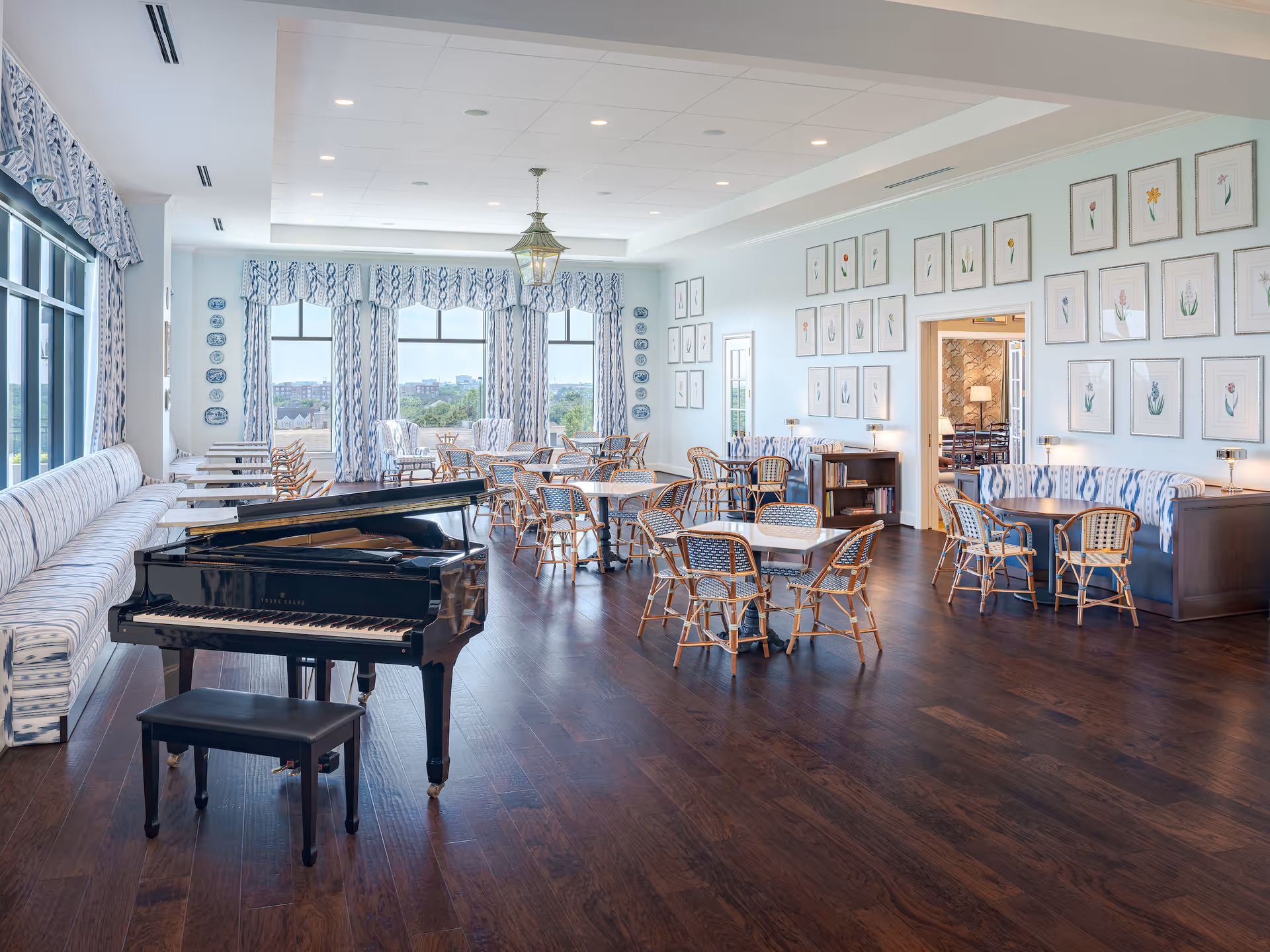 A spacious, well-lit dining room with multiple tables and chairs arranged throughout. A black grand piano with a matching bench is positioned on the left side near a long cushioned bench along the window wall. The room features large windows with patterned curtains, framed botanical prints on the walls, and dark hardwood flooring.