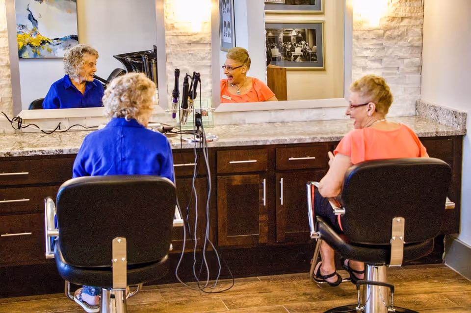 Two elderly women sitting in salon chairs facing a large mirror in a hair salon area. One woman is wearing a blue shirt and the other is wearing a coral shirt. They are smiling and appear to be engaged in a cheerful conversation. The salon has a granite countertop with hair styling tools and dark wooden cabinets underneath.