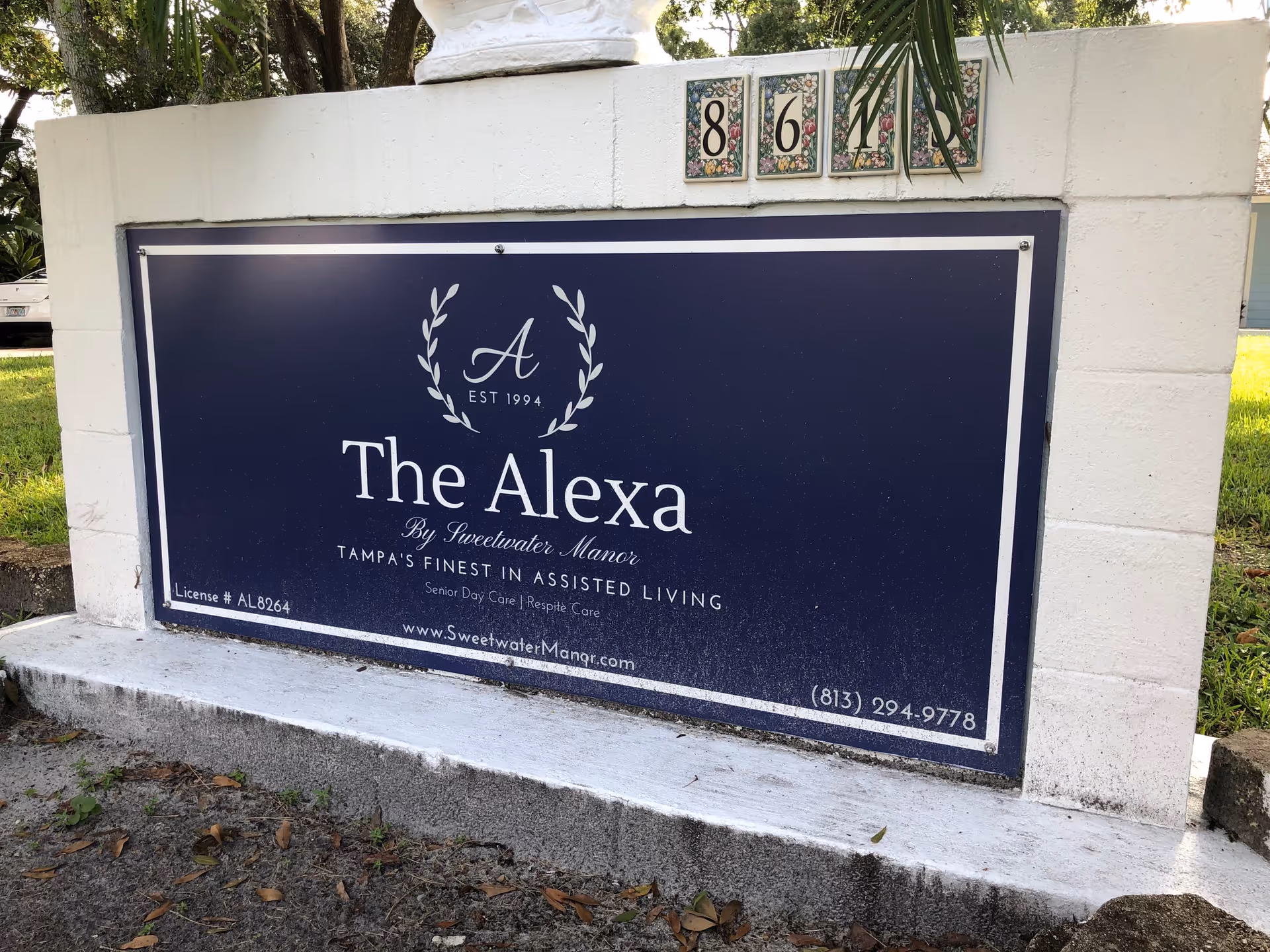 A blue and white sign mounted on a white concrete structure with the address number 861. The sign reads 'The Alexa By Sweetwater Manor, Tampa's Finest in Assisted Living, Senior Day Care | Respite Care' with contact information and a website URL.