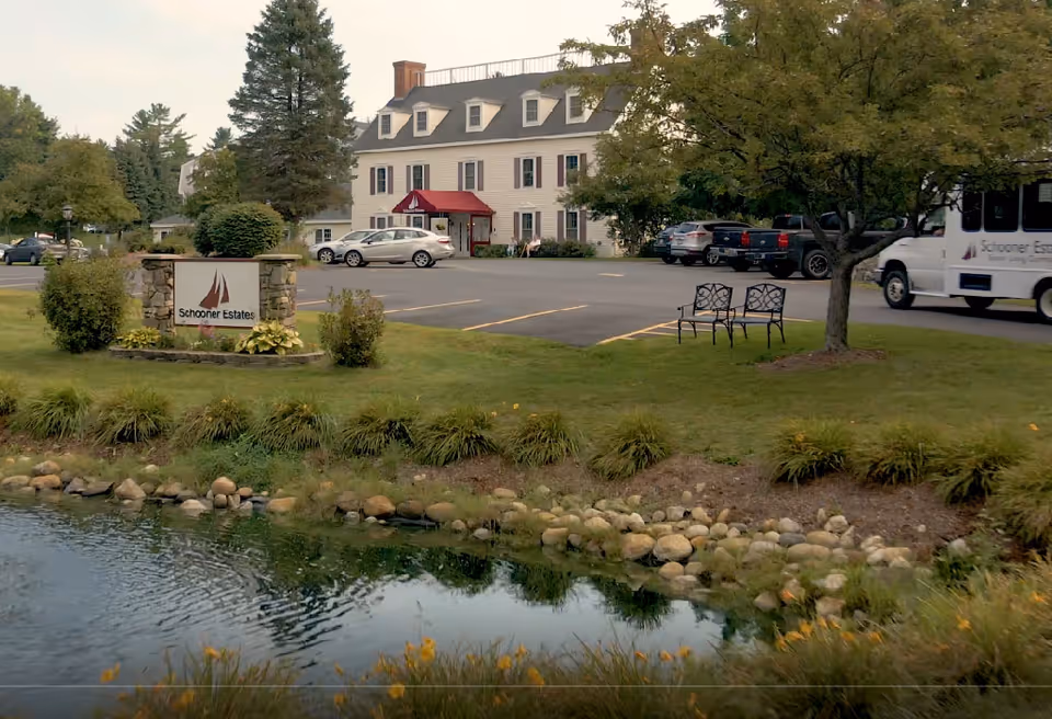 Exterior view of Schooner Estates facility with sign, benches, cars, and a pond.