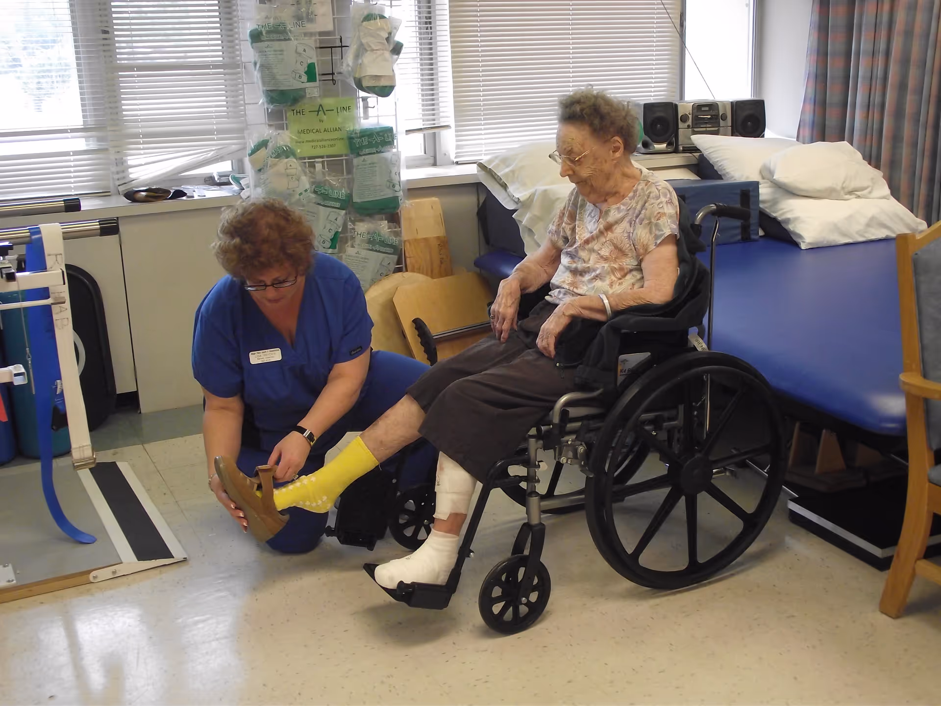 A healthcare worker in blue scrubs is helping an elderly woman in a wheelchair put on a shoe. The elderly woman has a bandaged foot and is wearing a yellow sock. They are in a medical rehabilitation room with exercise equipment, a padded treatment table, and windows with blinds in the background.