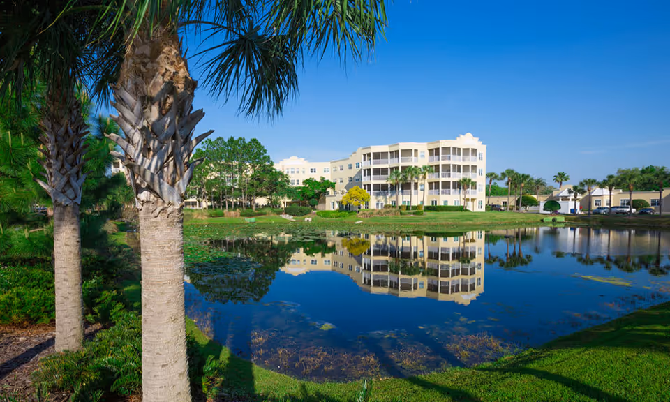 Multi-story senior living building reflected in a pond with palm trees in the foreground.