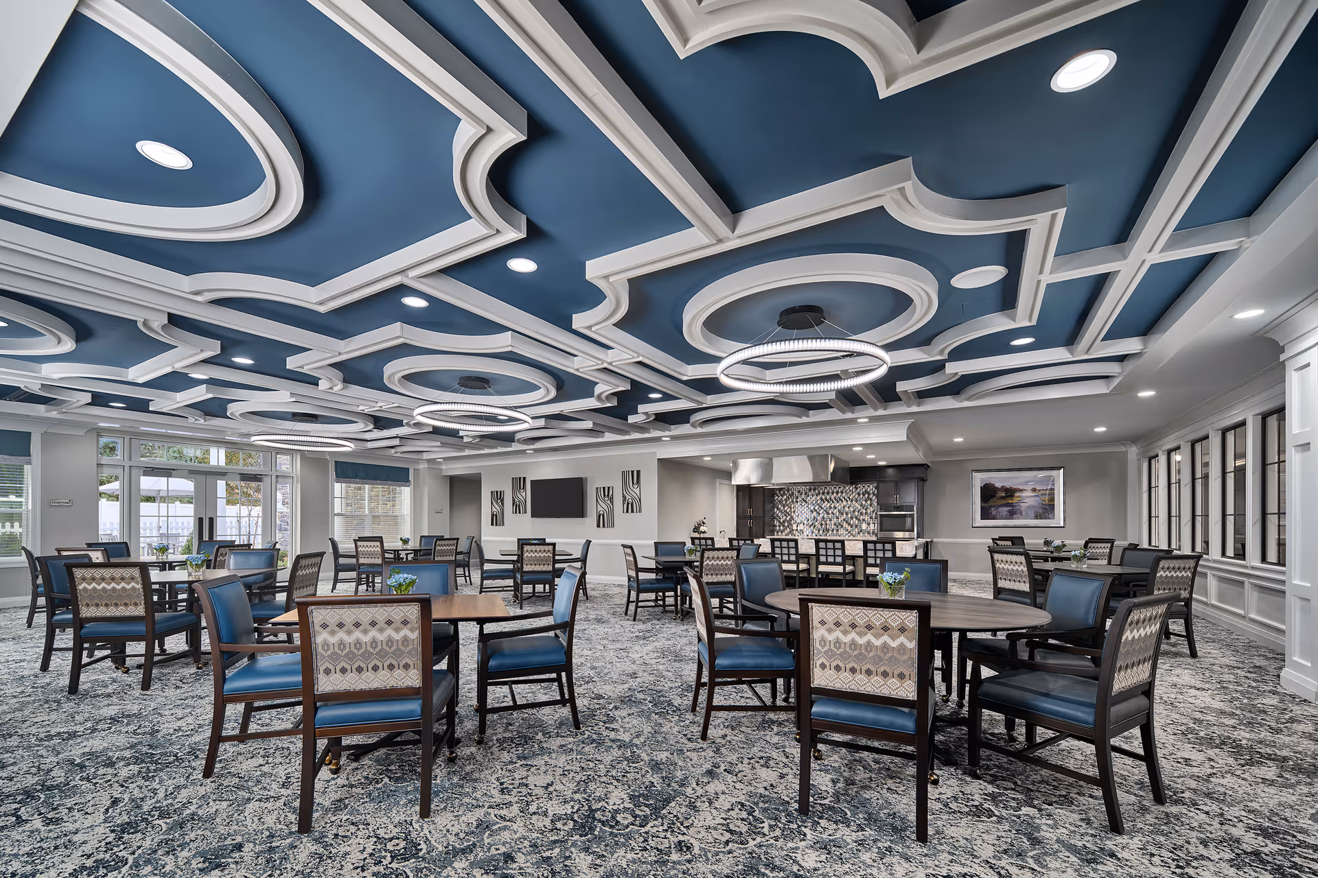 A spacious dining room with multiple round tables and chairs arranged neatly. The ceiling features an intricate blue and white geometric design with modern circular light fixtures. Large windows line one side of the room, allowing natural light to enter. The room has a patterned carpet and a kitchen area with a tiled backsplash and stainless steel appliances in the background.