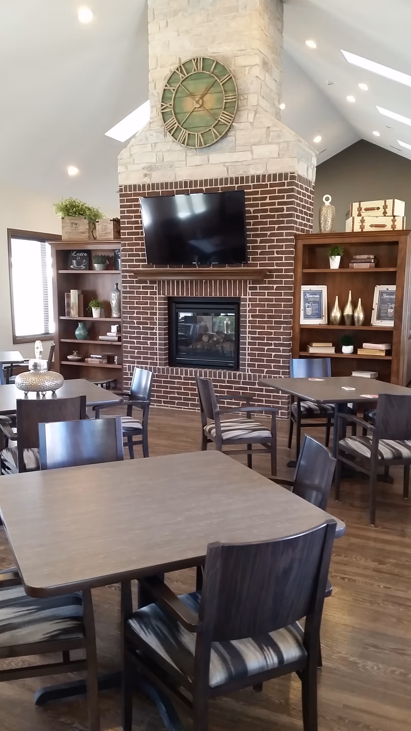 Interior view of a common area with several wooden tables and chairs arranged on a wooden floor. A brick fireplace with a mounted flat-screen TV above it is centered on the far wall. Above the fireplace is a large decorative clock. On either side of the fireplace are wooden bookshelves with decorative items and plants. The ceiling is vaulted with recessed lighting and skylights.