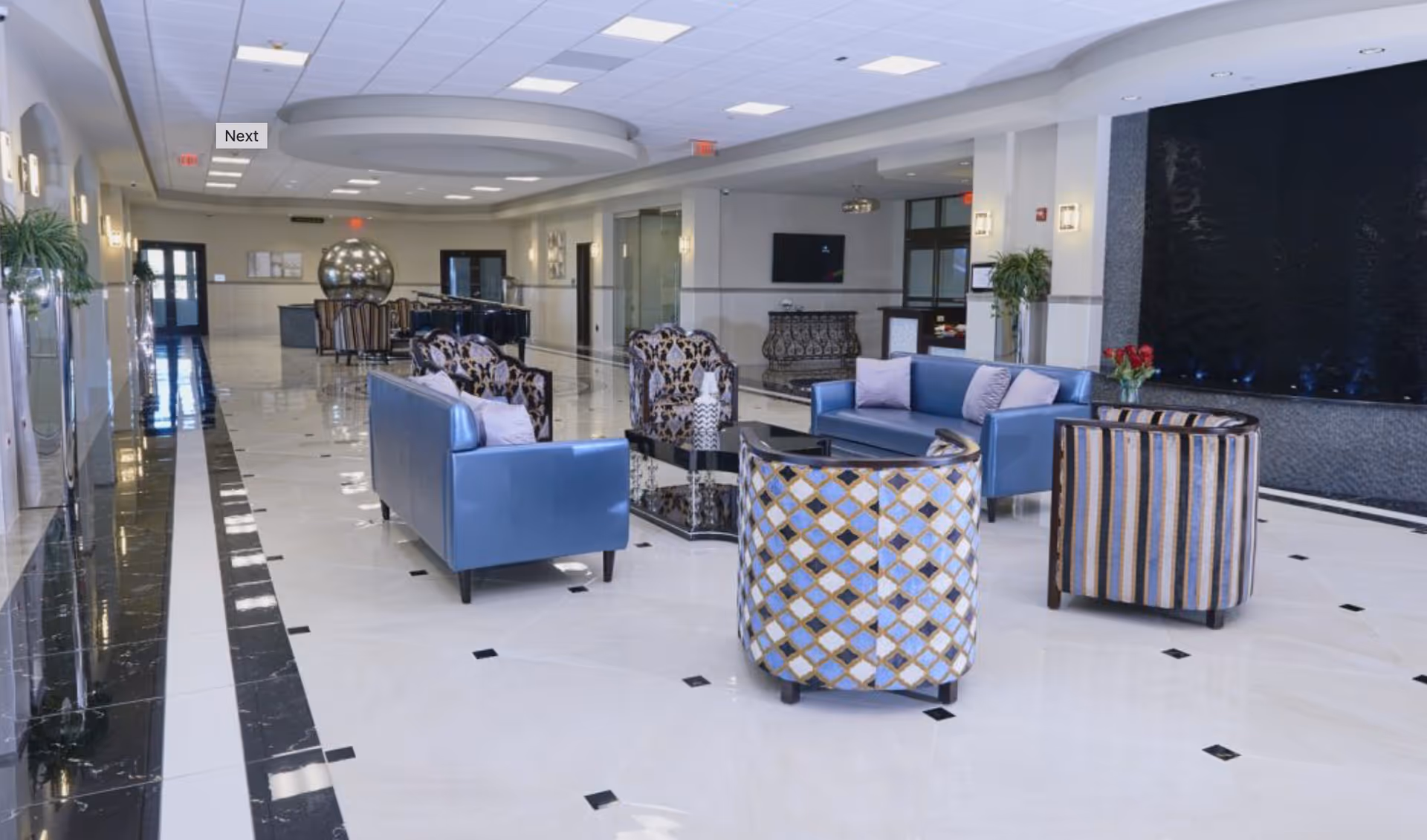 A spacious and modern lounge area with polished white and black tiled floors. The room features blue sofas, patterned armchairs, a black coffee table, and decorative plants. There is a large black water feature on the right wall and a television mounted on the far wall. The ceiling has recessed lighting and a circular architectural detail.