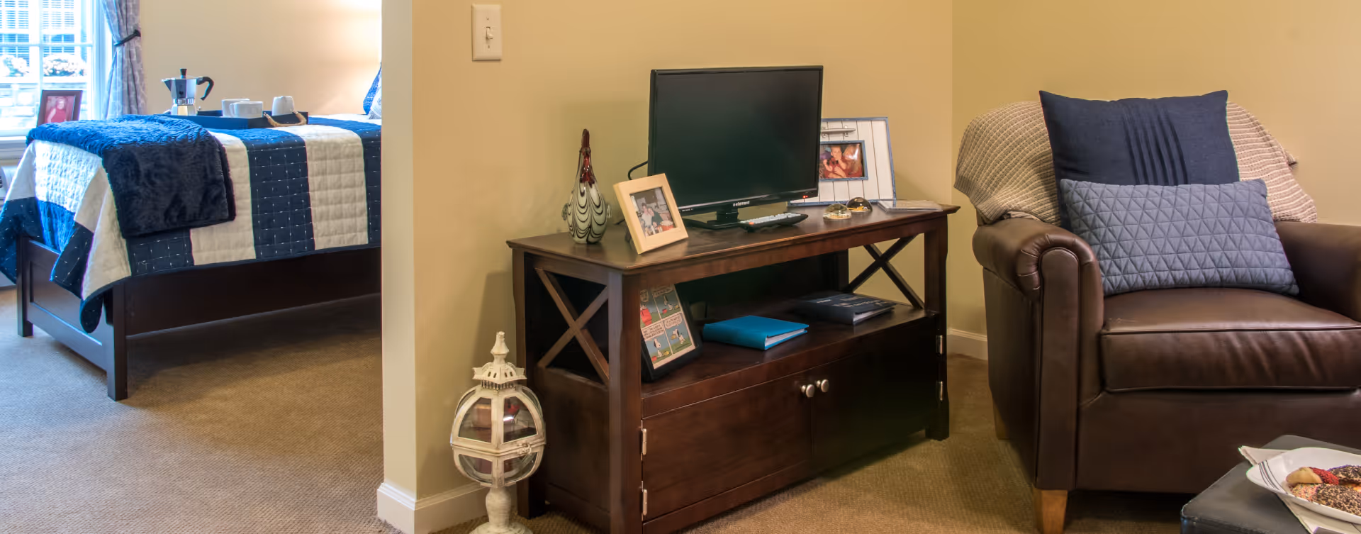 A cozy living space in a senior living facility featuring a brown leather armchair with two blue pillows and a beige throw blanket. Next to the armchair is a dark wooden TV stand with a flat-screen TV, framed photos, decorative items, and books. In the background, a bed with a blue and white quilt and a tray with a coffee pot and cups is visible near a window with curtains.
