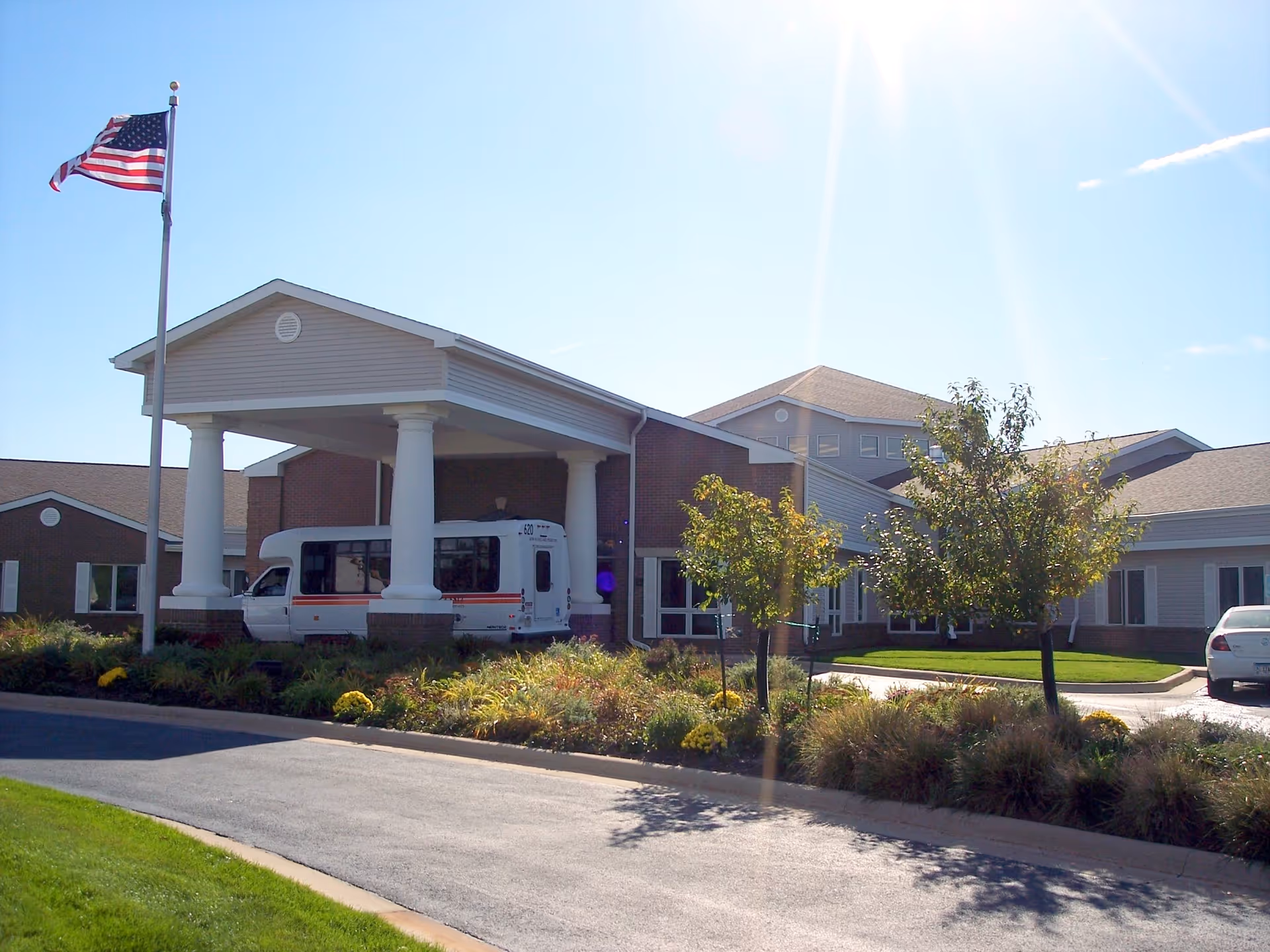 Exterior view of Lantern Park Specialty Care facility with a covered entrance supported by white columns, an American flag on a flagpole, a white shuttle bus parked under the entrance, landscaped greenery, and a clear sunny sky.