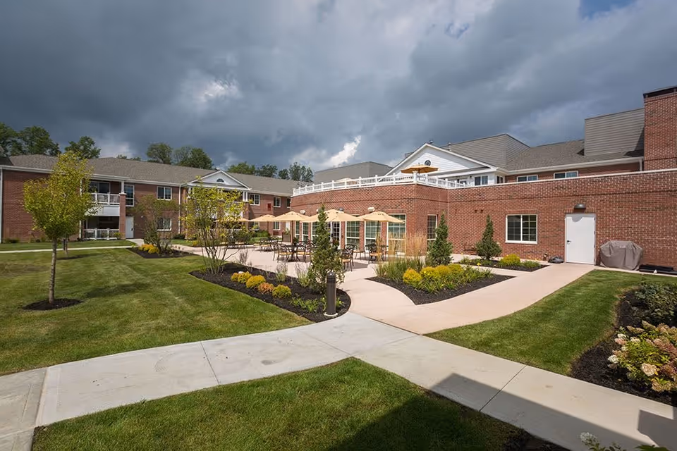 Outdoor courtyard area of a senior living facility with green lawns, young trees, landscaped flower beds, and a paved walkway. There are several tables with umbrellas on a patio adjacent to a red brick building under a cloudy sky.