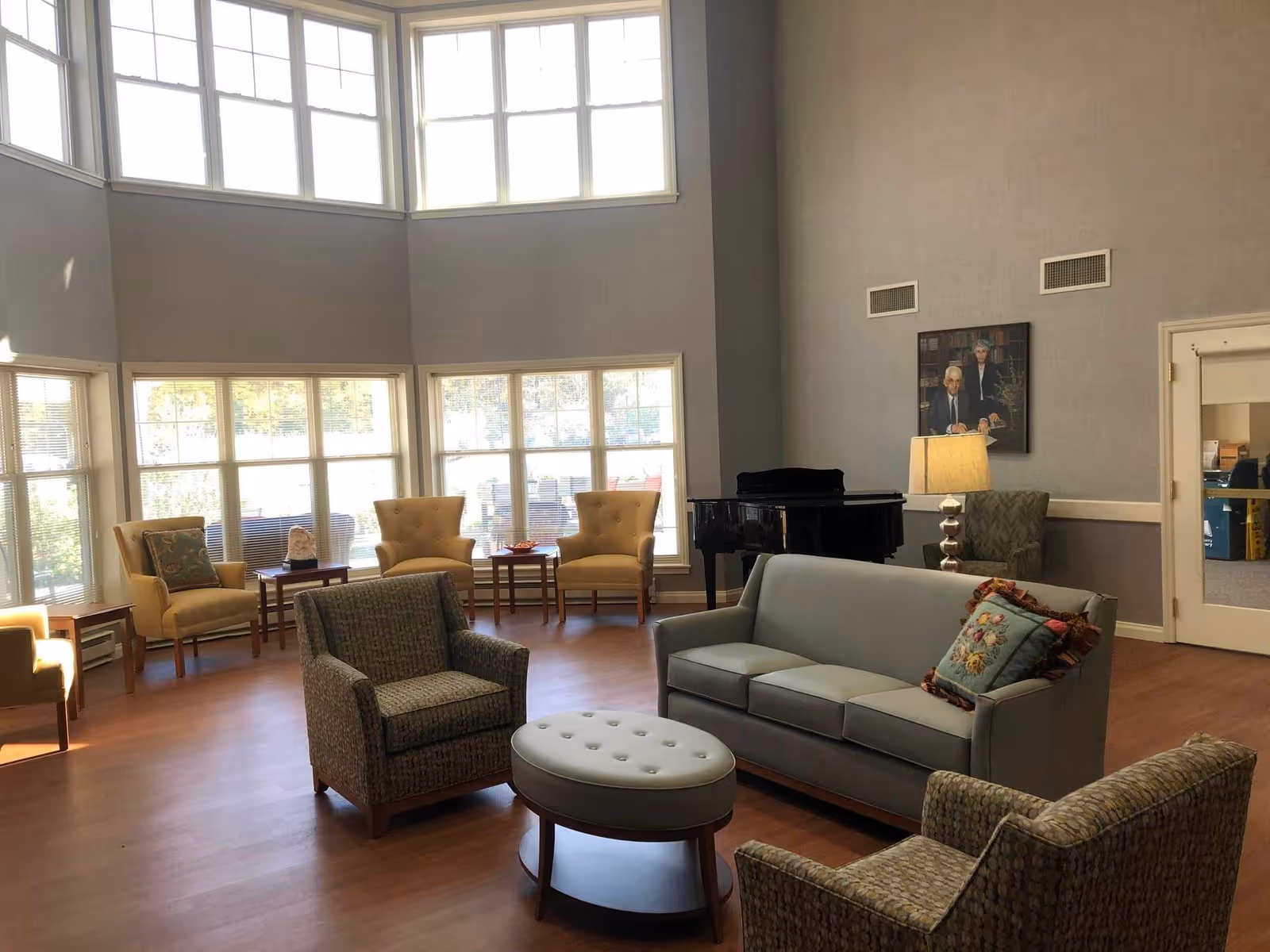 A spacious living room with large windows letting in natural light. The room features a gray sofa with a decorative pillow, two patterned armchairs, a round tufted ottoman, and several yellow armchairs arranged near the windows. A black grand piano is positioned against the wall, next to a floor lamp and a framed portrait. The walls are painted gray and the floor is wooden.