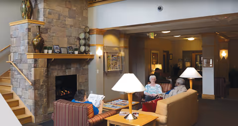 Seniors sitting and chatting in a cozy lounge area with sofas, table lamps, magazines, and a stone fireplace.