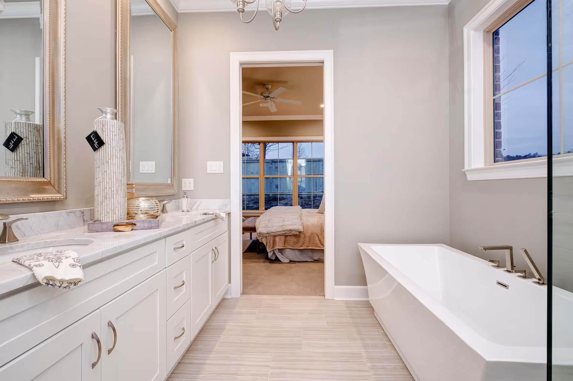 Modern bathroom with a white double-sink vanity on the left, a large rectangular bathtub on the right, and a doorway leading to a bedroom with a bed and ceiling fan visible. The bathroom has light-colored walls and floor tiles, with a large window above the bathtub.