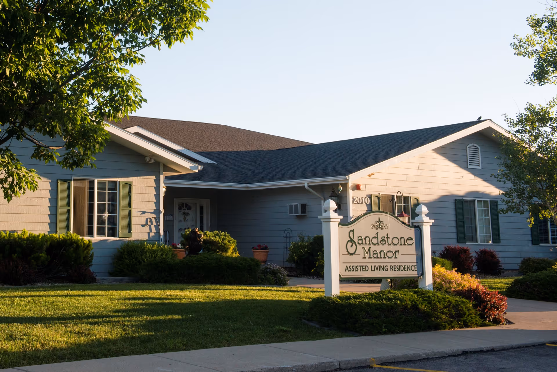 Exterior view of Sandstone Manor, an assisted living residence, showing a single-story building with gray siding, green shutters, and a sign in front surrounded by well-maintained landscaping and trees.