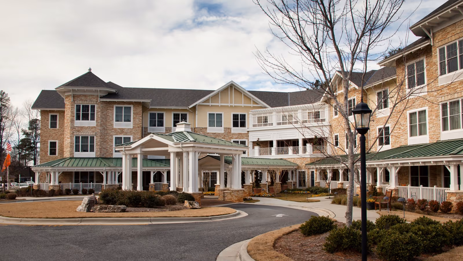 Exterior view of a multi-story senior living facility building with beige and stone facade, green metal roofs, a covered entrance with white pillars, landscaped bushes, and a curved driveway under a partly cloudy sky.