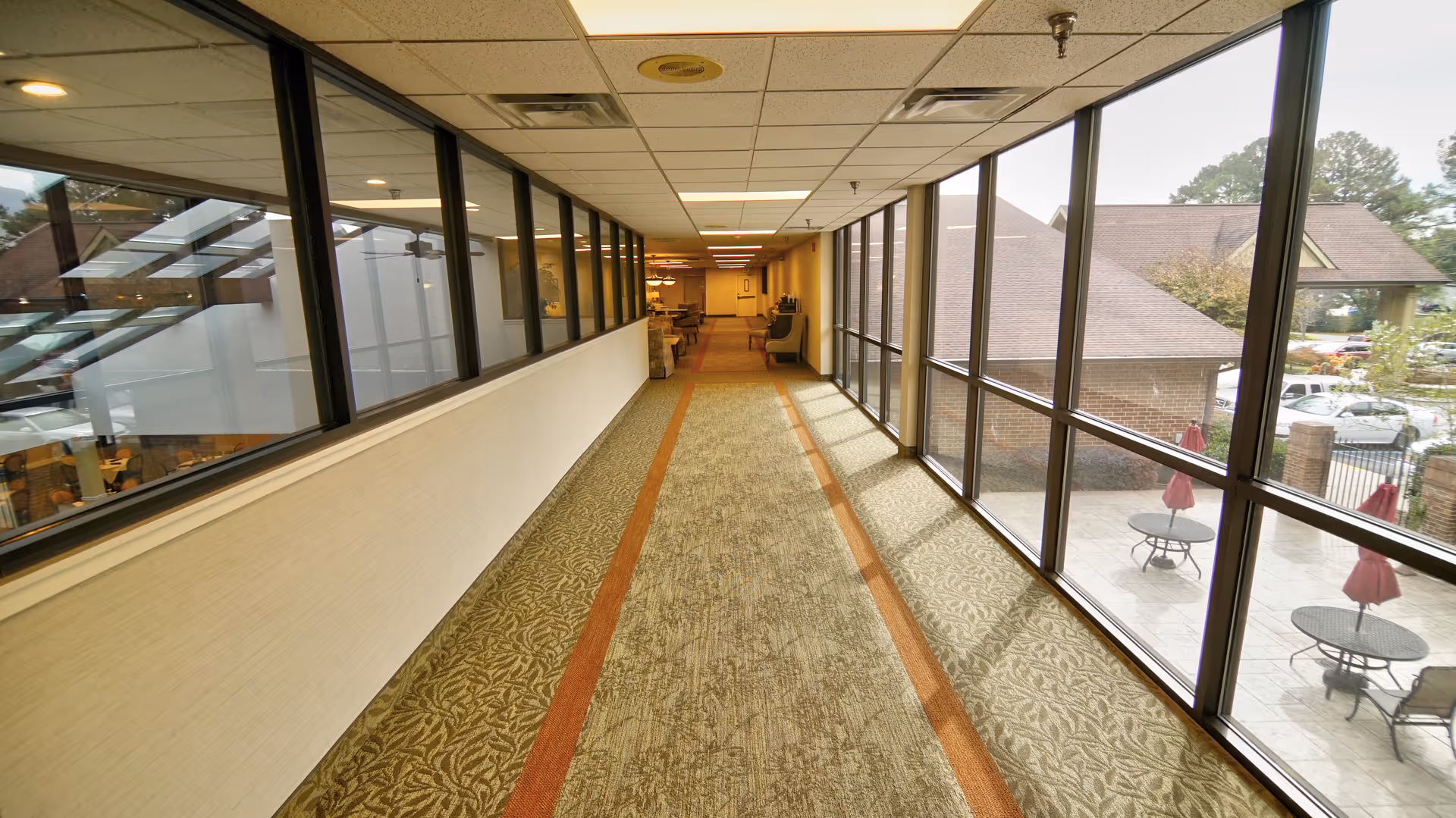 A long indoor hallway with large windows on the right side showing an outdoor patio area with tables and red umbrellas. The left side has a wall with windows looking into another room. The hallway is carpeted with a patterned design and has ceiling lights.