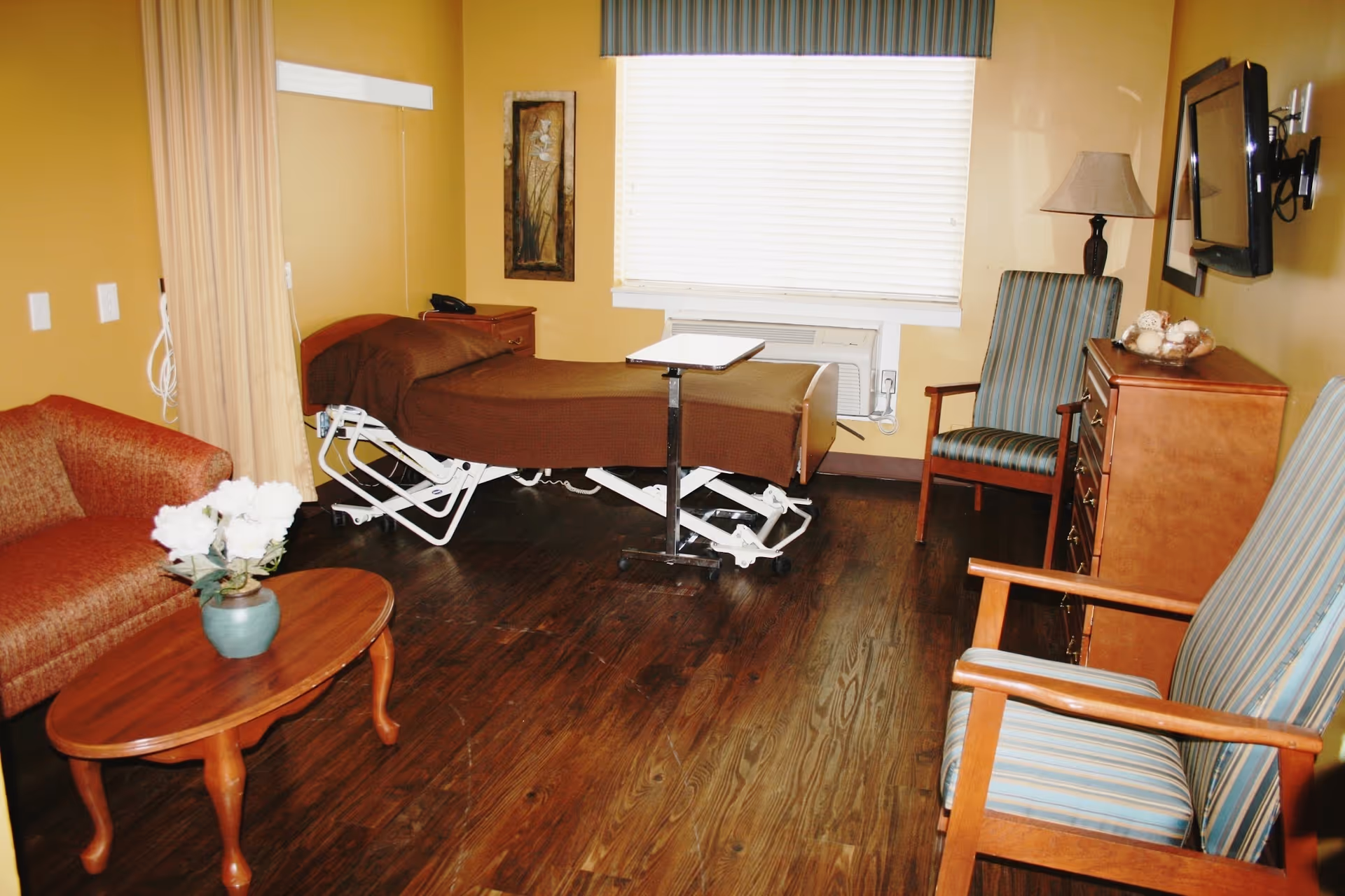 A rehabilitation center room with a hospital bed covered in a brown blanket, a small adjustable overbed table, a wooden dresser with a lamp and decorative bowl, two striped armchairs, a brown couch, a wooden coffee table with a vase of white flowers, and a window with blinds and a striped valance.