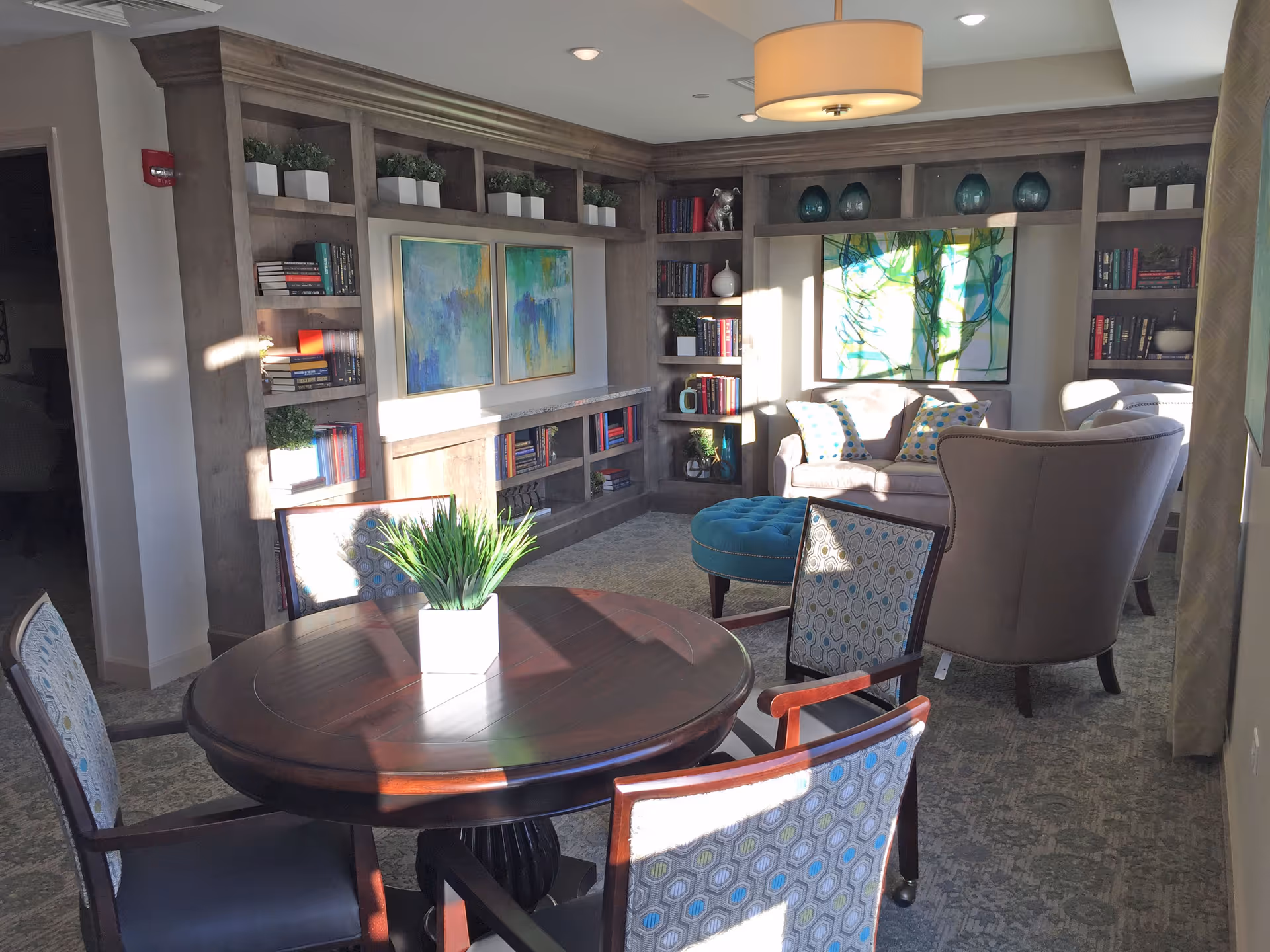 A cozy living room area with a round wooden table and four patterned chairs in the foreground. Behind the table is a built-in wooden bookshelf filled with books, plants, and decorative items. There is a beige sofa with patterned pillows, a blue tufted ottoman, and two beige armchairs. The room is well-lit with natural light coming through a window, and a modern ceiling light fixture hangs above.