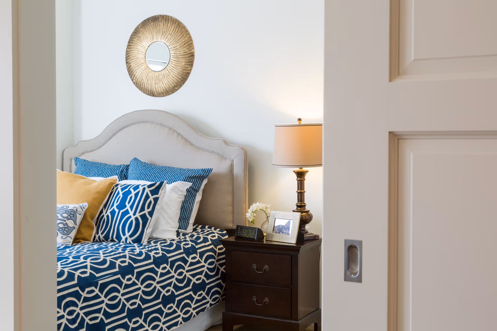 View into a bedroom with a beige upholstered headboard, blue patterned bedding and pillows, and a nightstand with a lamp and framed photo.