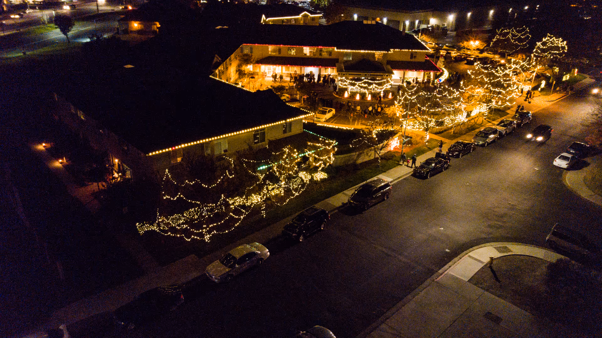A nighttime aerial view of a building decorated with numerous white string lights outlining the roof, trees, and bushes. Several cars are parked along the street adjacent to the building, and people are gathered near the entrance and outdoor area illuminated by the lights.