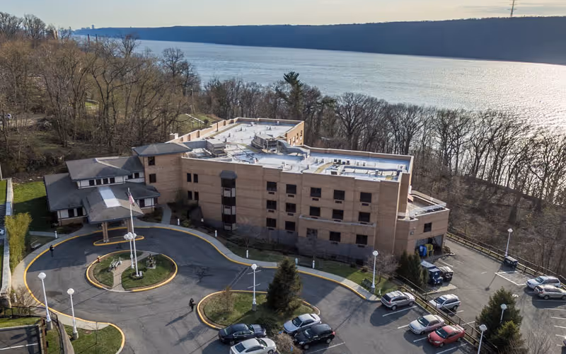 Aerial view of a multi-story brick building surrounded by a circular driveway and parking lot with several cars. The building is located near a large body of water with trees and hills in the background under a clear sky.