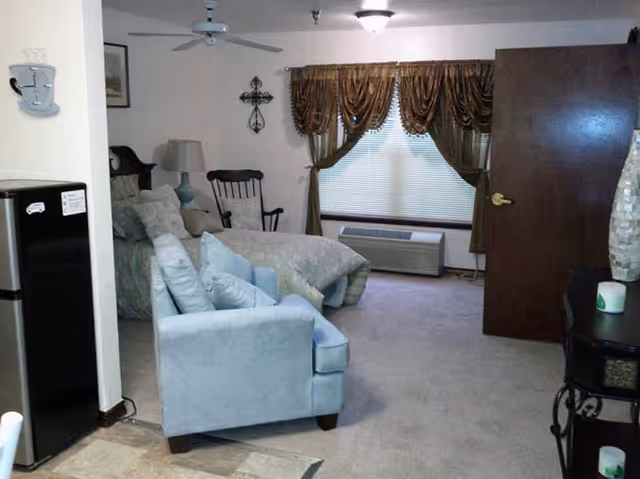 Interior view of a senior living bedroom with a bed, a blue armchair, a rocking chair, a lamp, a window with brown curtains, and an air conditioning unit below the window. The door to the room is open, and part of a small refrigerator is visible on the left side.
