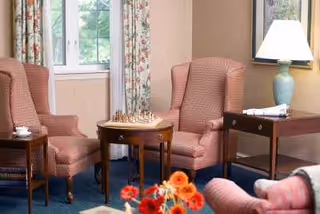 A cozy sitting area with two upholstered armchairs facing each other across a small wooden table with a chessboard on it. There is a window with floral curtains in the background, a side table with a lamp and a telephone, and a vase with orange and red flowers in the foreground.