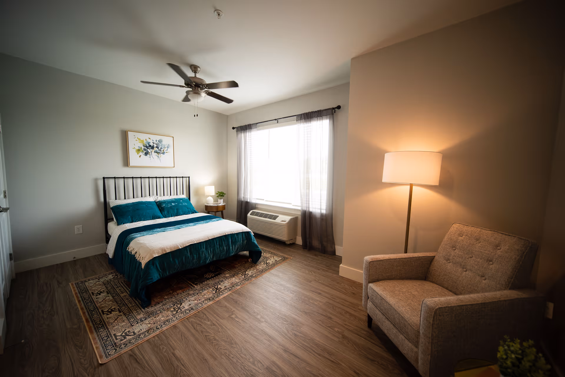 Bright furnished bedroom with a bed, bedside table, window AC unit, armchair, floor lamp, ceiling fan and area rug.