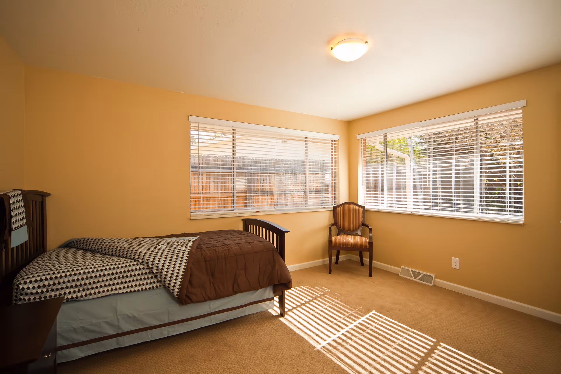 A bright bedroom with beige walls and carpet, featuring a single bed with a brown and patterned blanket, a wooden nightstand, and a striped upholstered chair near two large windows with white blinds letting in sunlight.