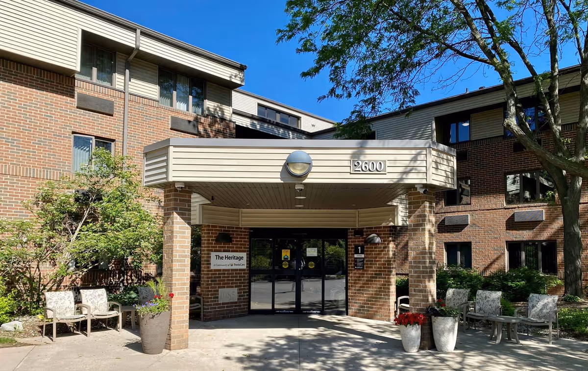 Entrance of a brick building with a covered porch displaying the address number 2600. There are chairs and potted plants on either side of the entrance. A sign next to the door reads 'The Heritage'. Trees and shrubs surround the area under a clear blue sky.