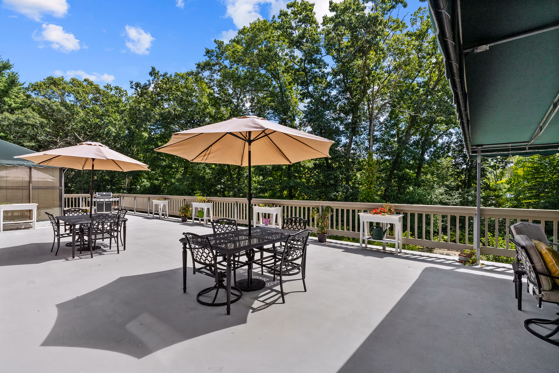 Outdoor patio area with metal tables and chairs under beige umbrellas, surrounded by a wooden railing and lush green trees under a blue sky with some clouds.