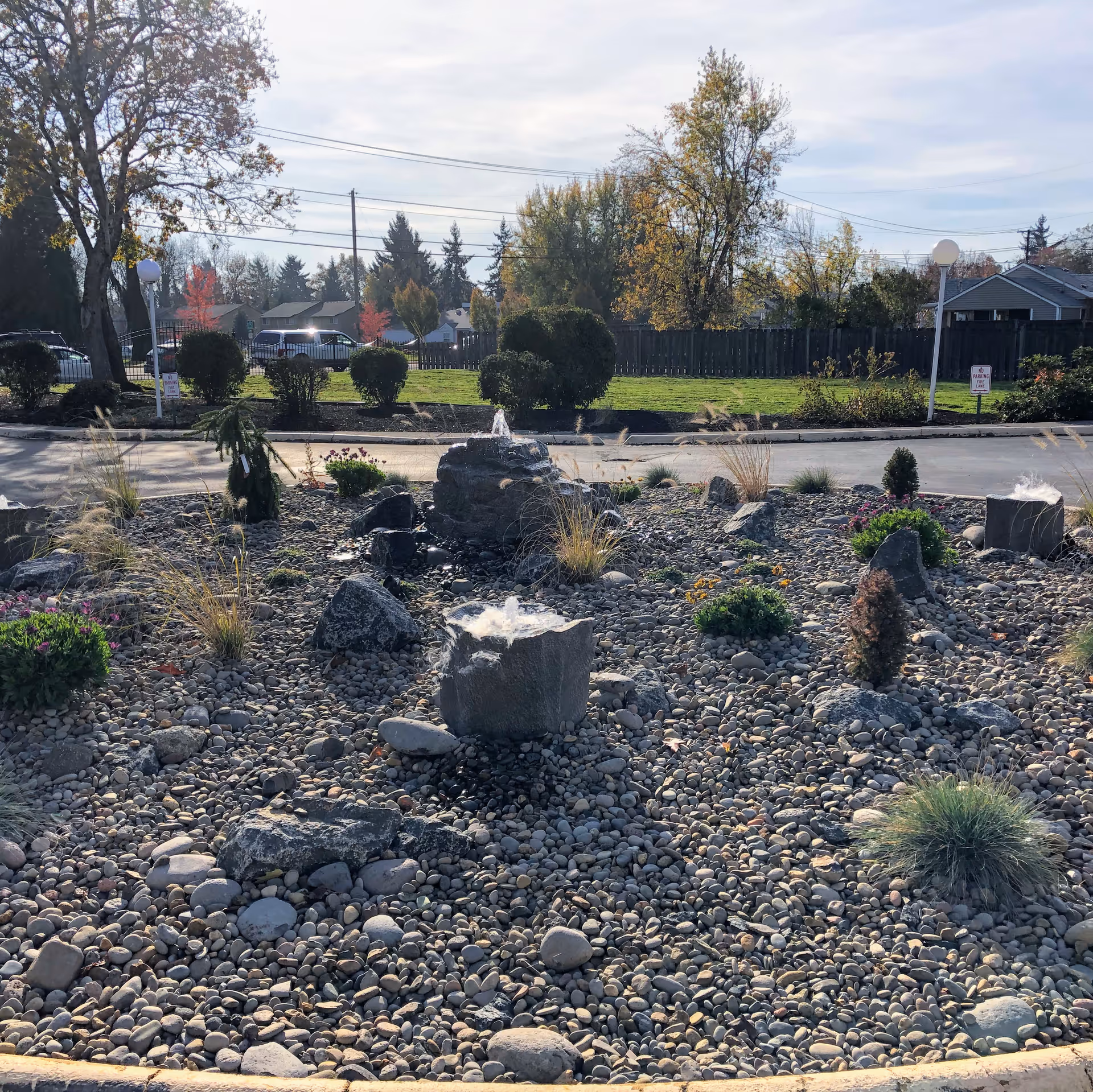 Rock and pebble landscaped courtyard featuring small stone fountains, ornamental grasses, and shrubs with a driveway, lawn, and trees in the background.
