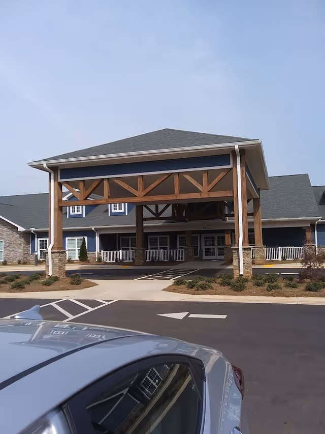 Front exterior view of Davie Nursing & Rehabilitation Center showing a large covered entrance with wooden beams and stone pillars, a paved driveway with parking spaces, and part of a silver car in the foreground.