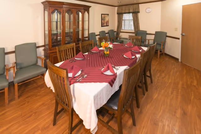 A dining room with a long rectangular table covered with a white tablecloth and a burgundy table runner. The table is set with folded burgundy napkins, silverware, and a small flower arrangement in the center. Surrounding the table are wooden chairs with dark cushions. In the background, there is a wooden china cabinet with glass doors, several upholstered chairs along the walls, a window with curtains, and a clock on the wall.