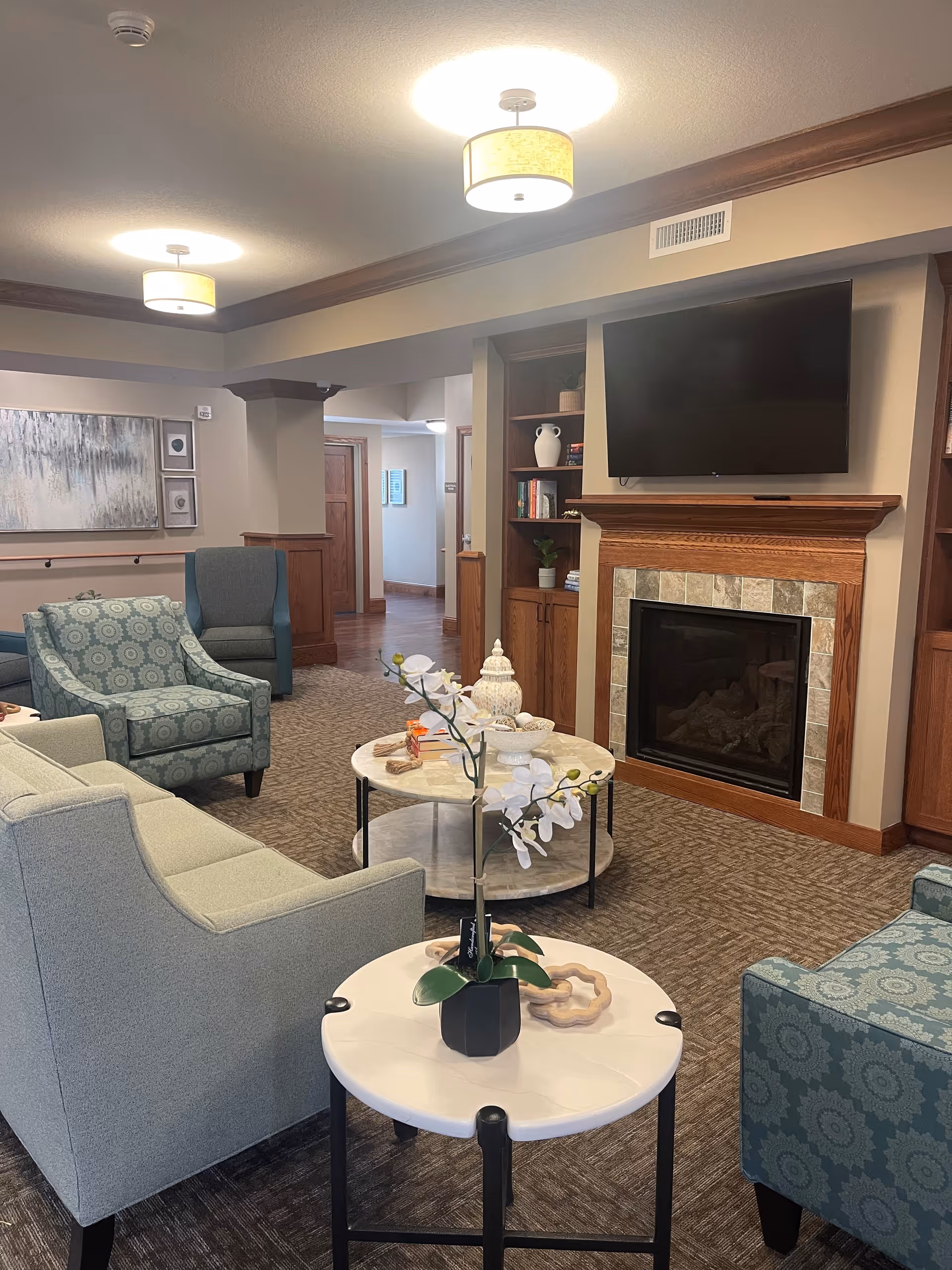 A cozy senior living facility lounge area with comfortable upholstered chairs and sofas arranged around a round coffee table and a smaller side table with a potted orchid. The room features a fireplace with a mounted flat-screen TV above it, built-in wooden shelves with decorative items and books, and soft overhead lighting. The carpeted floor and neutral wall colors create a warm and inviting atmosphere.