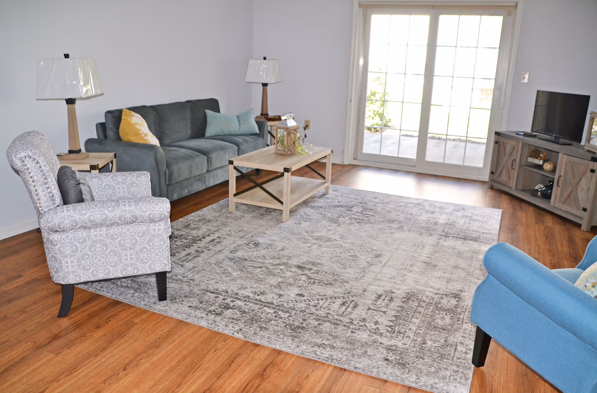 A bright living room with wooden flooring, a large gray patterned area rug, a gray sofa with colorful pillows, a patterned armchair, a blue armchair, two wooden side tables with lamps, a wooden coffee table with a decorative lantern, and a TV on a wooden stand near sliding glass doors leading outside.