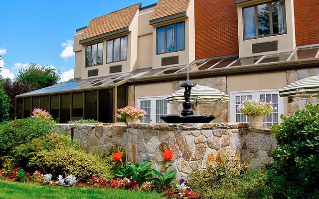 Outdoor view of a building with stone walls and large windows. There is a black water fountain in the center surrounded by flower pots with pink flowers and green plants. The area is landscaped with bushes, flowers, and a green lawn under a blue sky with some clouds.