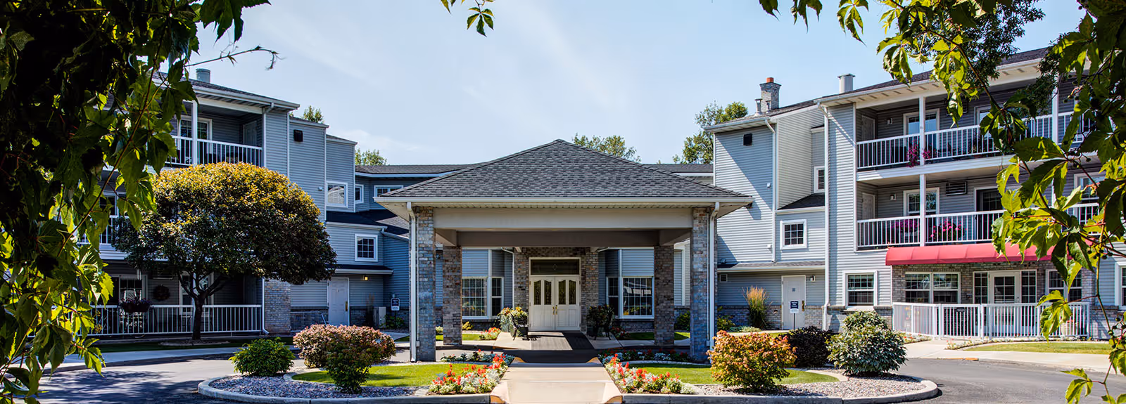 Front exterior view of The Village Senior Residence showing a three-story building with balconies, a covered entrance, landscaped garden beds with flowers and shrubs, and trees framing the scene under a clear blue sky.