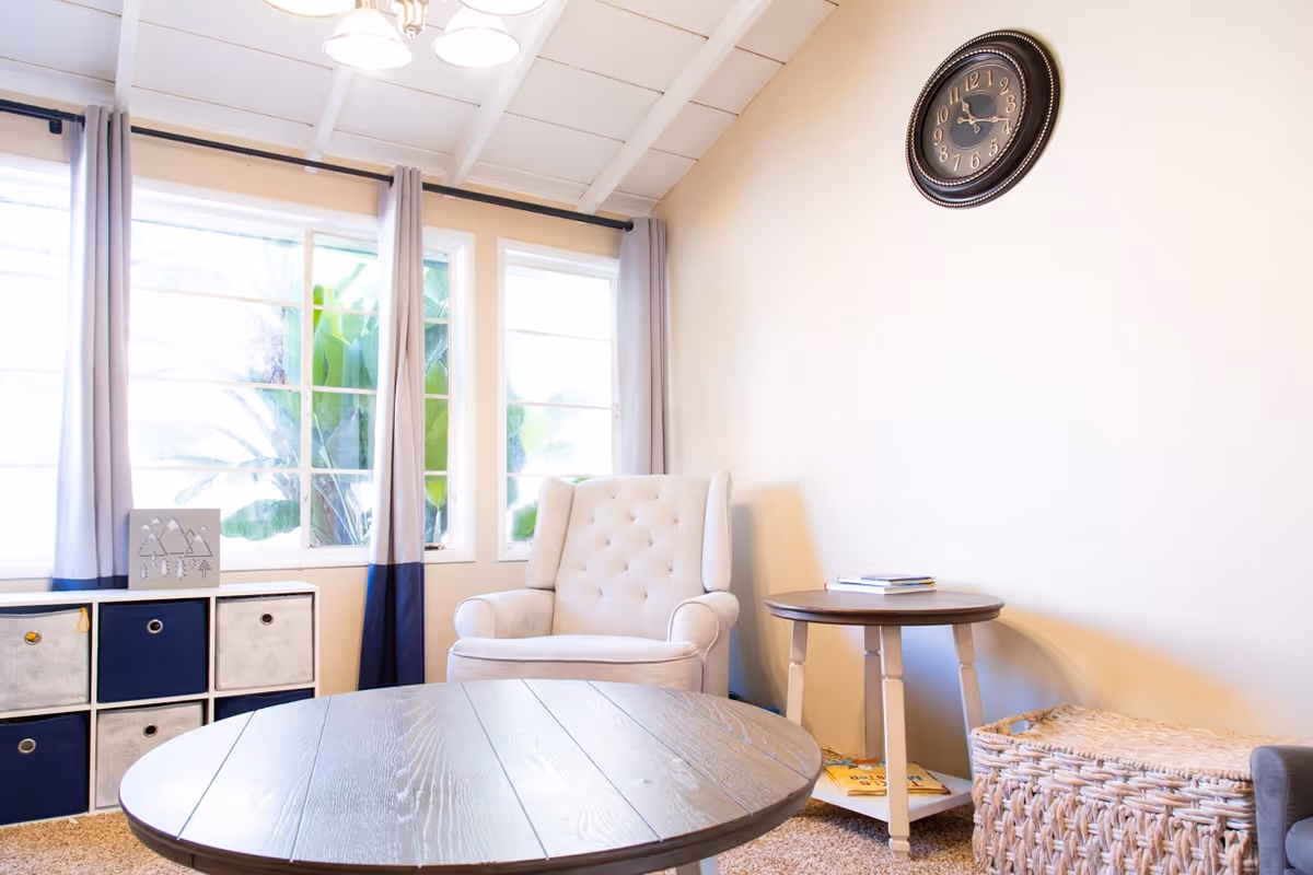 A cozy living room with a beige tufted armchair, a round wooden coffee table, a small side table with books, a woven basket, and a wall clock. Large windows with curtains allow natural light to fill the room.