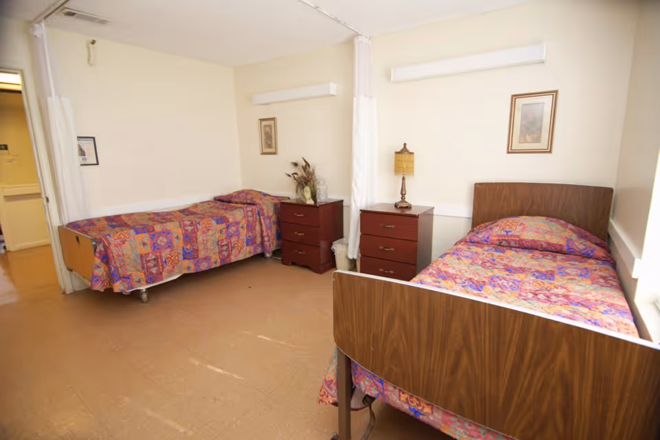 A simple nursing facility bedroom with two single beds, each covered with colorful patterned bedspreads. Between the beds are two wooden nightstands, one with a lamp and the other with a decorative vase. The room has beige walls and a linoleum floor, with a curtain partially drawn near the doorway.