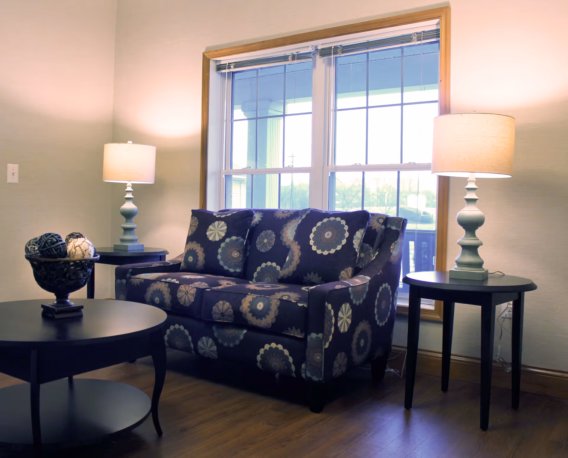 A cozy living room area with a patterned loveseat featuring circular floral designs, flanked by two side tables each holding a lamp with a beige shade. A large window with wooden trim lets in natural light, and a round coffee table with decorative balls in a bowl is in the foreground. The room has wooden flooring and light-colored walls.