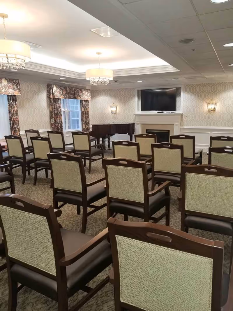 A well-lit room with multiple rows of wooden chairs with cushioned seats and backs arranged facing a fireplace with a mounted flat-screen TV above it. There is a grand piano in the corner, patterned wallpaper, floral curtains on the windows, and elegant ceiling lights.