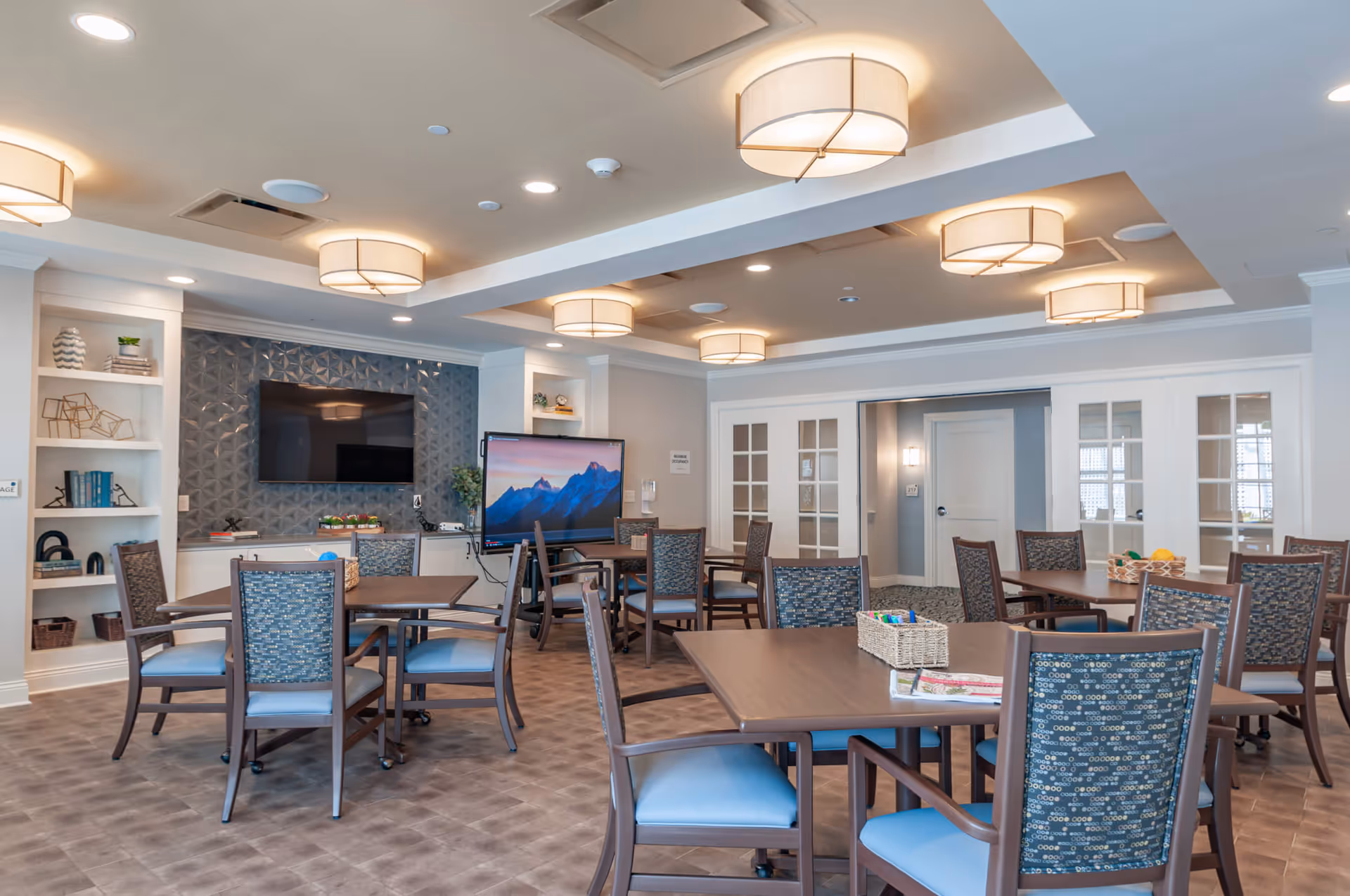 Bright communal dining/activity room with multiple tables and chairs, a wall-mounted TV, shelving, and overhead drum-style lights.