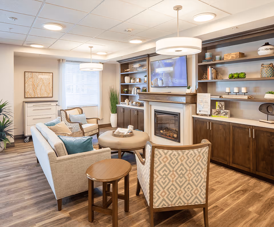 A cozy living room area in Sonata Vero Beach featuring a beige sofa with blue and brown cushions, two patterned armchairs, a round ottoman, and a small wooden side table. The room has wood flooring, built-in dark wood shelves with decorative items, a fireplace with a mounted TV above it, and two modern pendant lights hanging from the ceiling. A window with white blinds and light curtains lets in natural light.