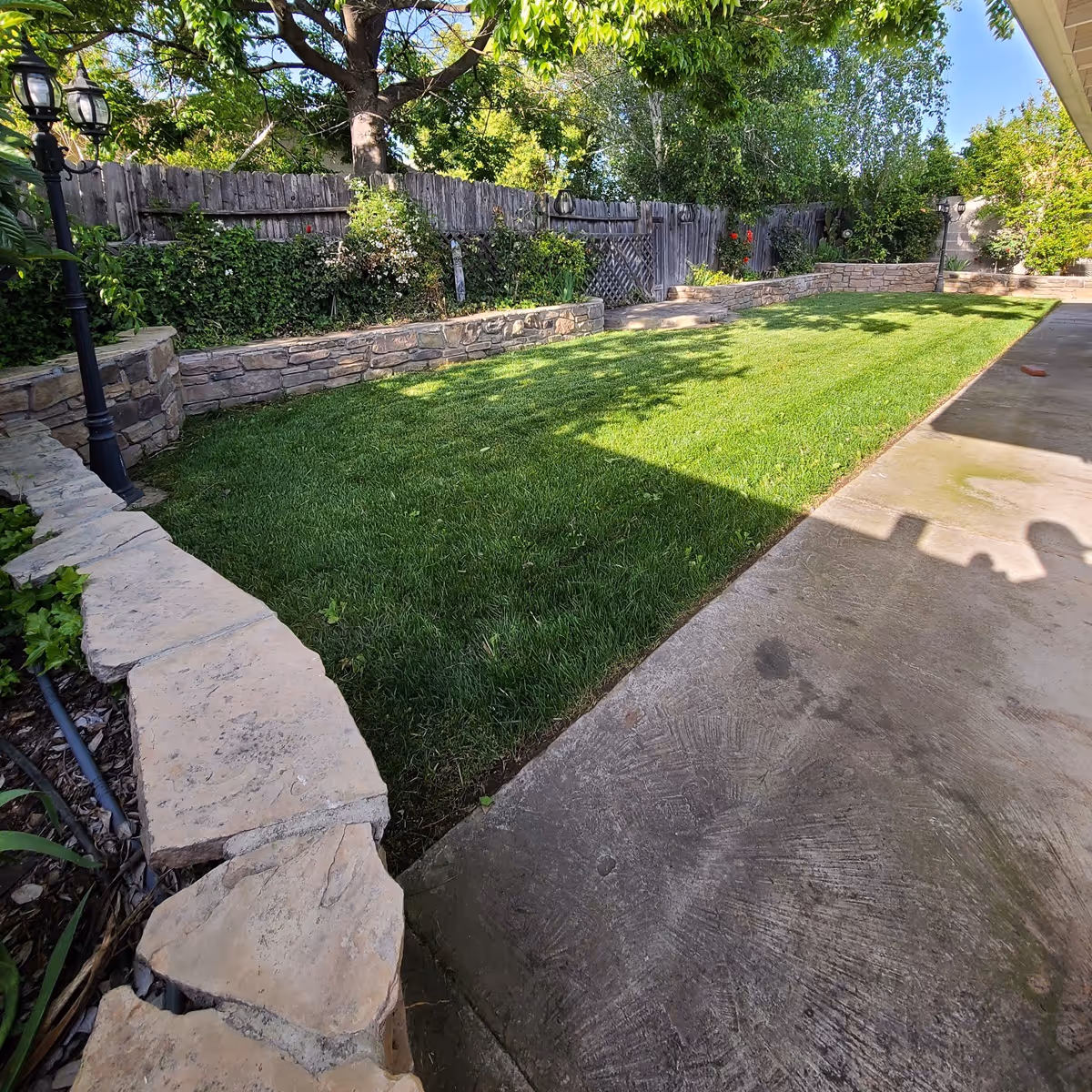Backyard with green lawn, stone walls, and a concrete patio along a wooden fence.