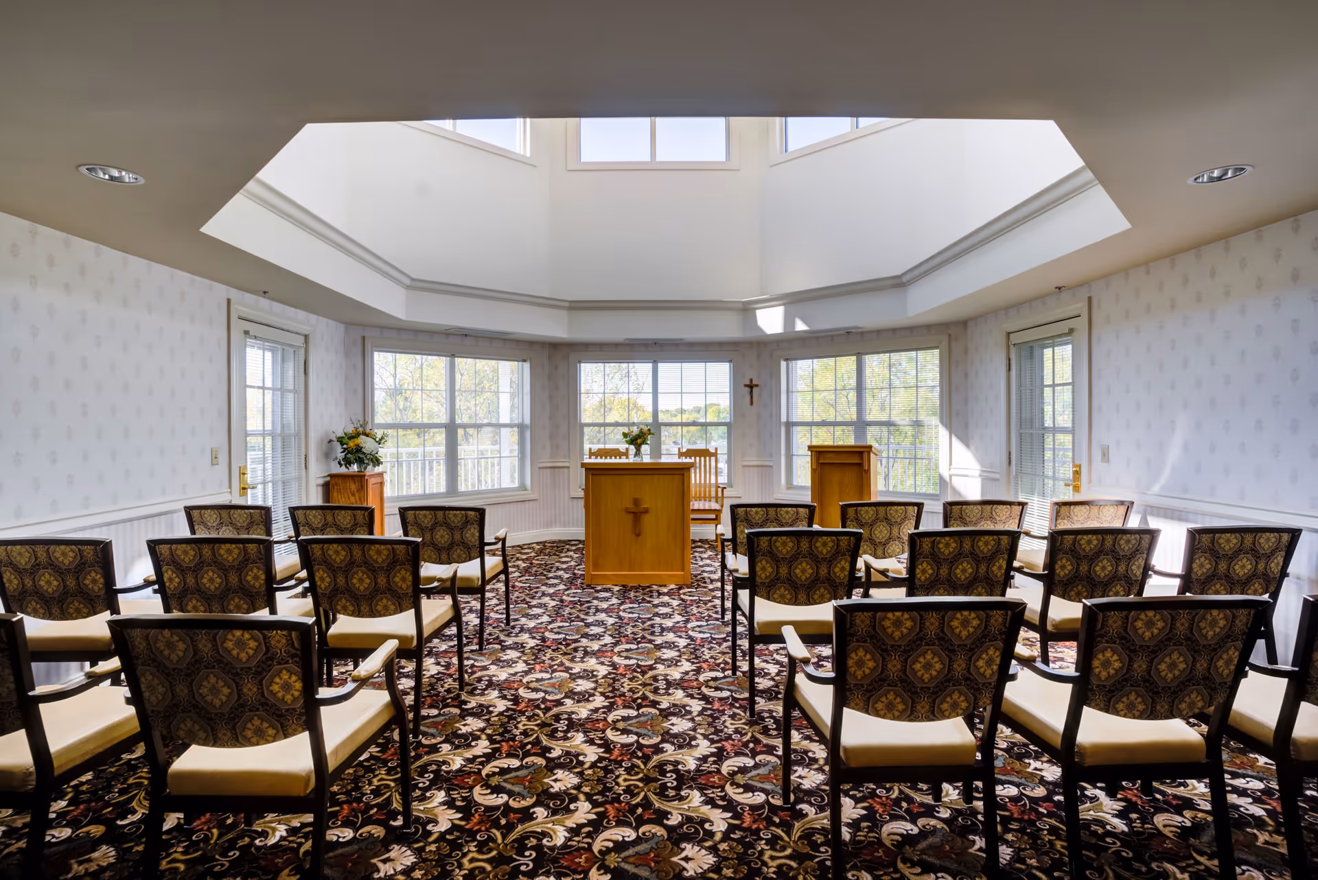A small chapel or meeting room with rows of patterned chairs facing a wooden podium and lectern, both adorned with crosses. The room has large windows letting in natural light and a patterned carpet with floral designs.