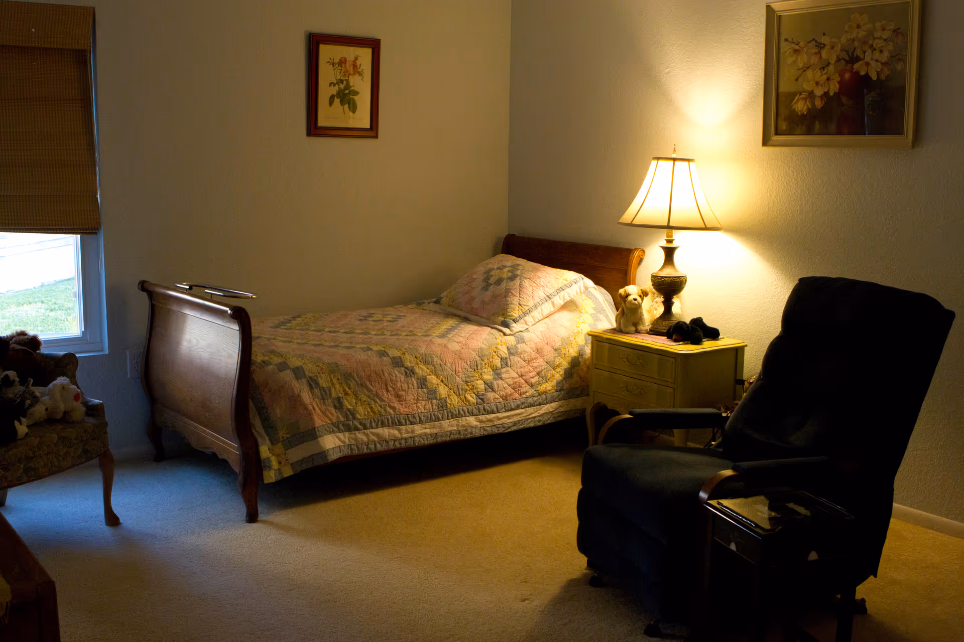 A cozy bedroom with a wooden bed covered in a pastel-colored quilt. A nightstand beside the bed holds a lamp and two stuffed animals. There is a dark cushioned armchair next to the nightstand. The room has light-colored walls with two framed floral paintings and a window with a bamboo shade partially drawn.