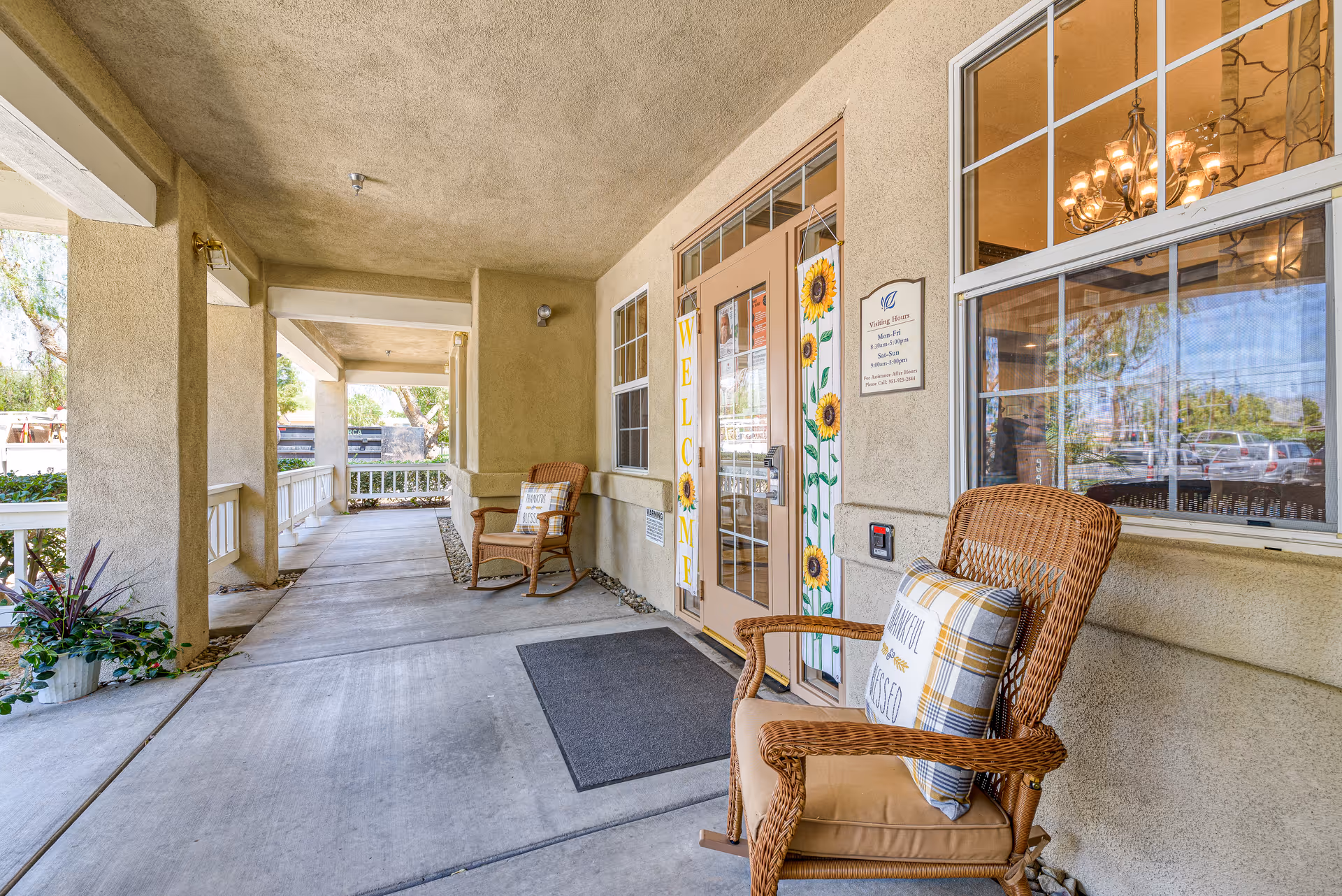 Covered outdoor porch area at the entrance of the Cottages at Hemet facility featuring two wicker rocking chairs with cushions, a welcome sign with sunflowers hanging on the door, a doormat, and a window reflecting a chandelier and parked cars outside.